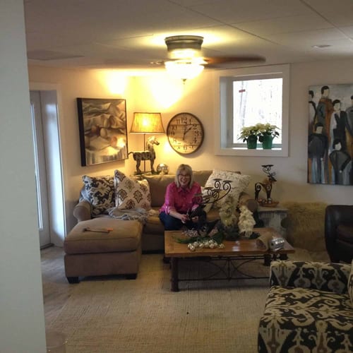 Relaxed woman sitting on a beige sectional sofa in a cozy living room, decorated with contemporary artwork, warm lighting, and festive holiday decor, emphasizing home renovation and interior design services.